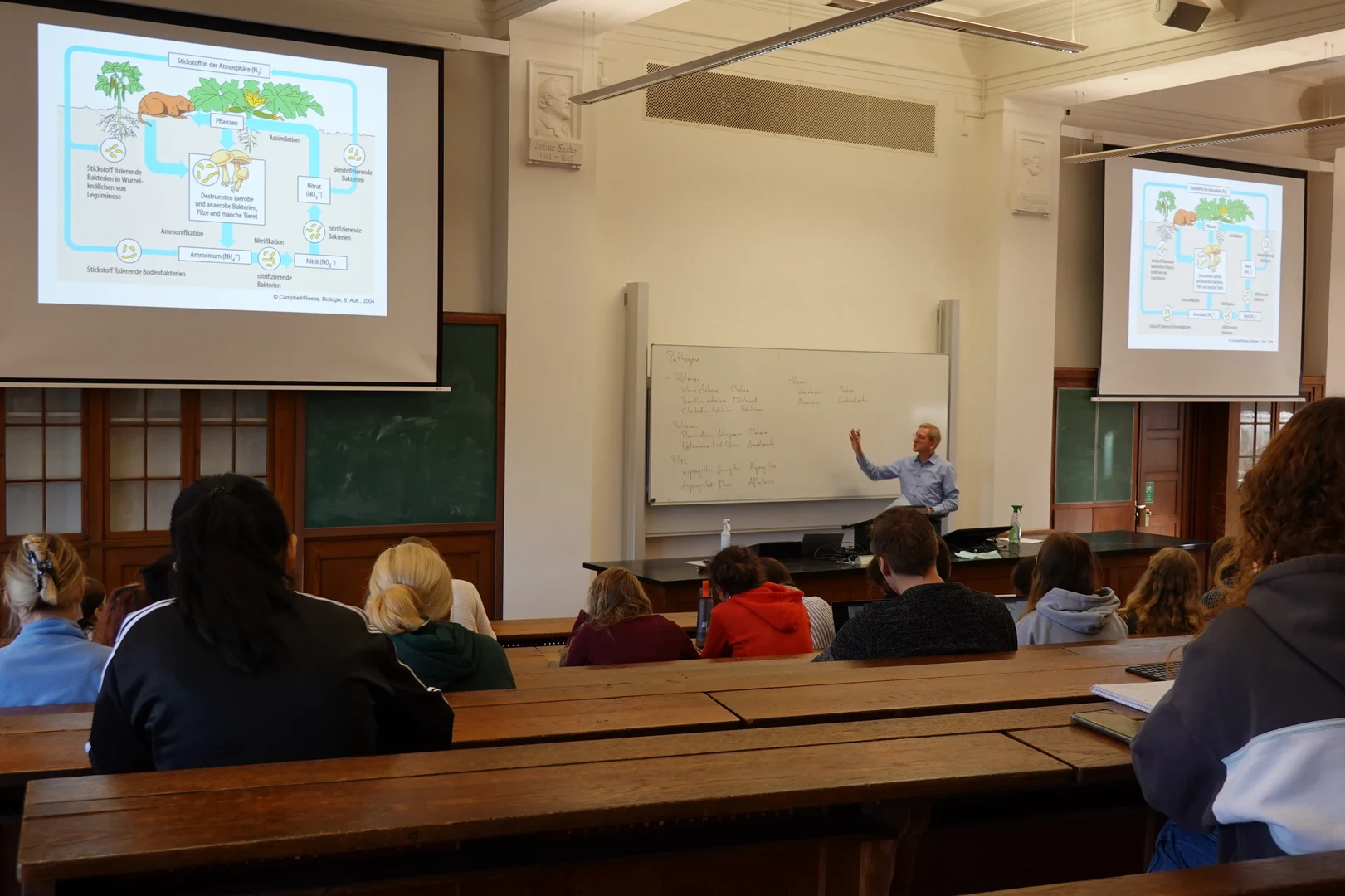 Eine Wissenschaftlerin und ein Wissenschaftler arbeiten hinter einer Glasfassade und mischen Chemikalien mit Großgeräten.